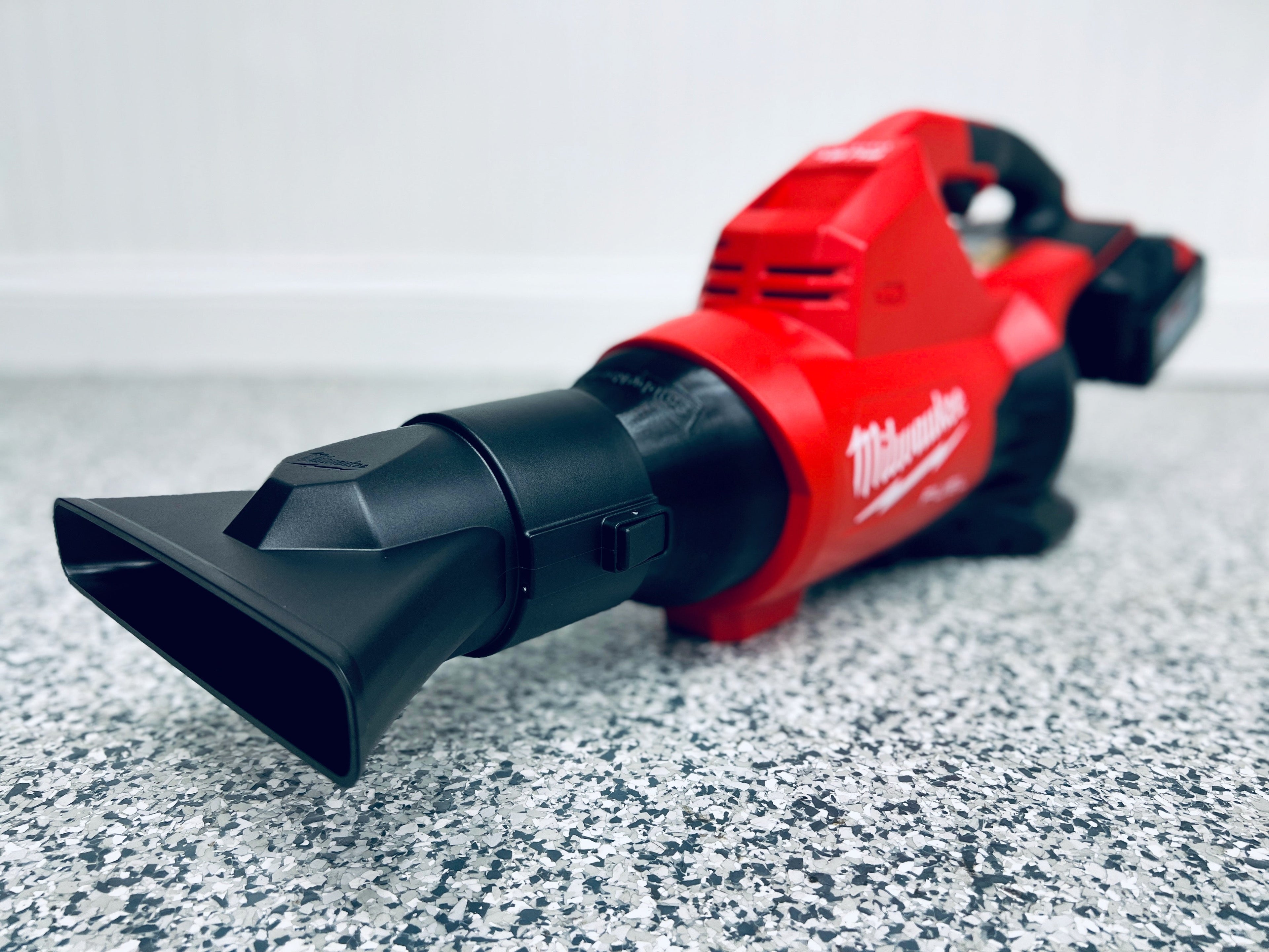 A red Milwaukee M18 FUEL Dual Battery Leaf Blower (2824-20) with a wide black nozzle is on a gray speckled floor, highlighting its compatibility with the STUBBY® Nozzle Adapter designed for this model.