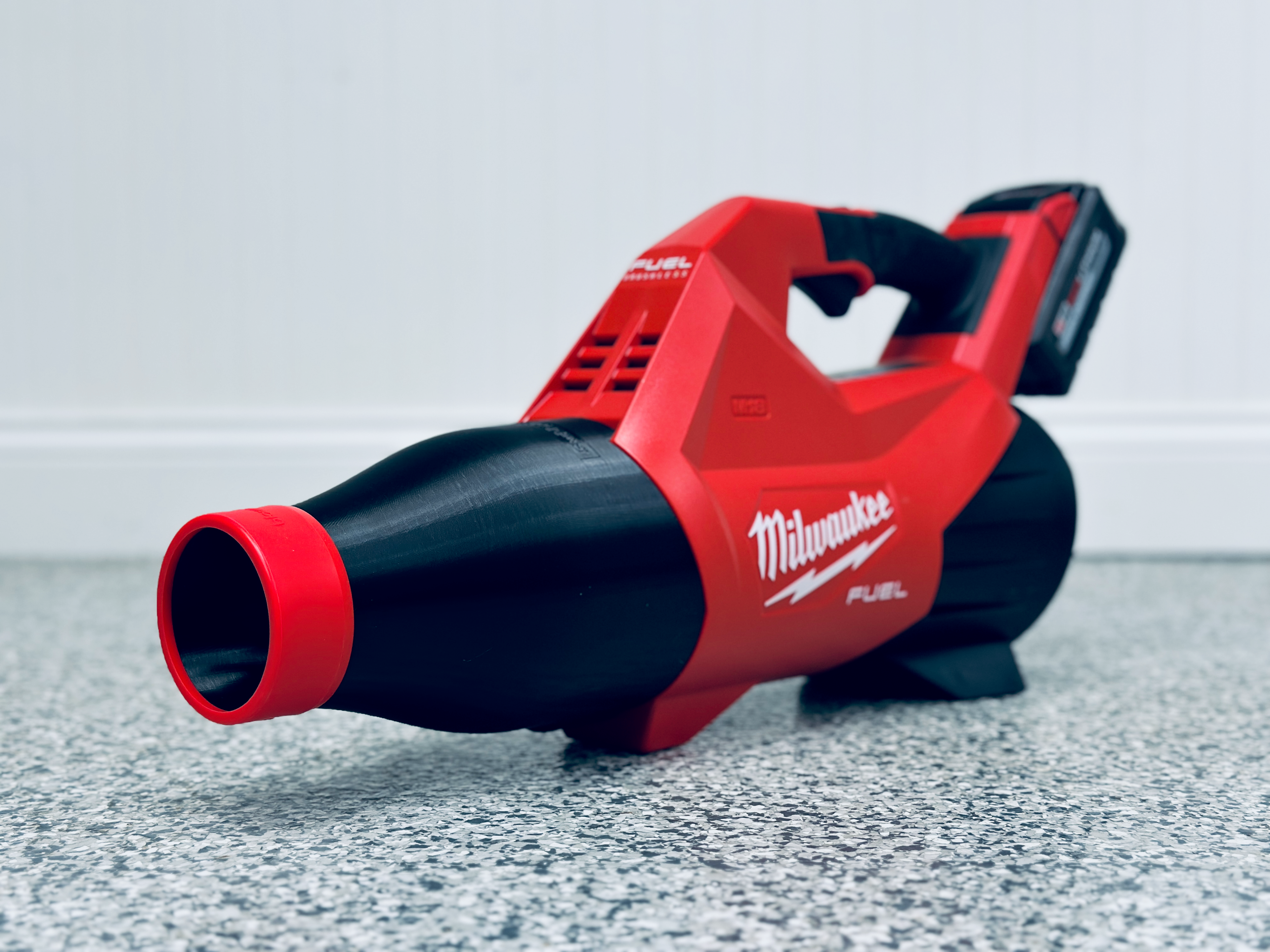 A STUBBY® Car Drying Nozzle for Milwaukee M18 FUEL Gen III Single Battery Leaf Blower (3017-20) fitted on a red and black blower with a silicone tip rests on a speckled gray floor against a white paneled wall, nozzle facing forward.