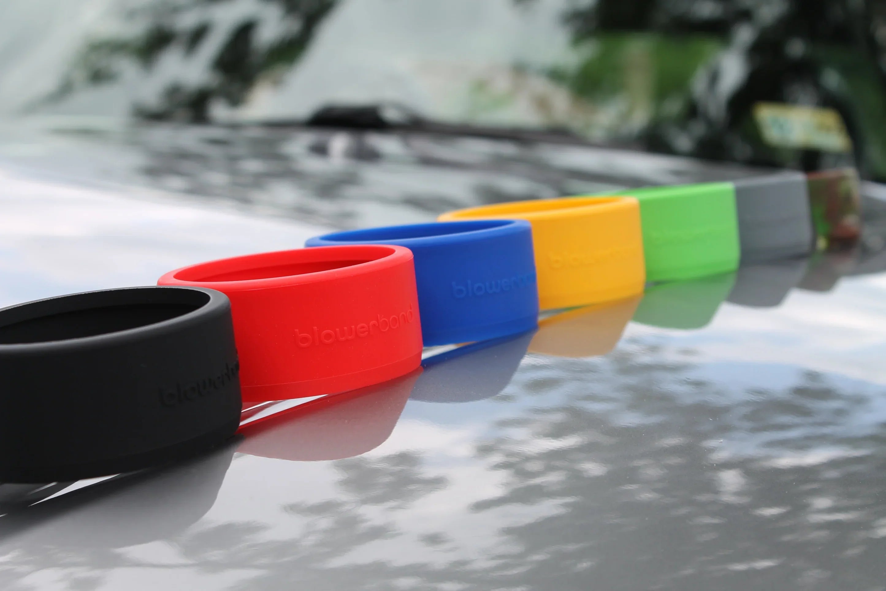 Six Blowerband® Guards for STUBBY Car Drying Nozzles—black, red, blue, yellow, green, and gray—are lined up diagonally on a reflective surface with blurred trees behind. Each provides colorful silicone protection for your nozzle.