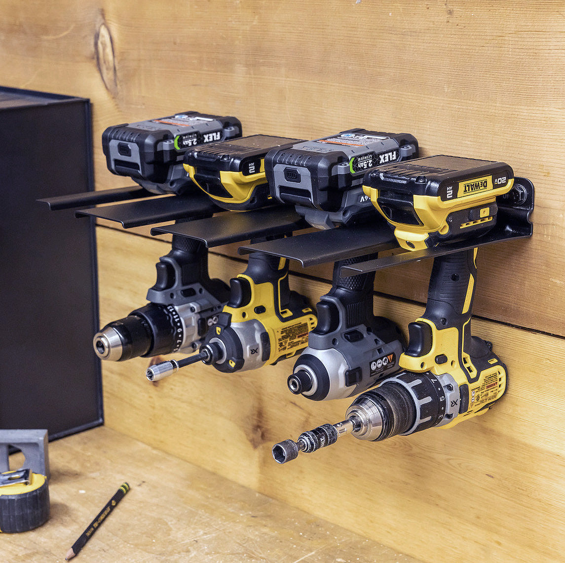 Three yellow and black cordless power drills hang from the E-Drill Power Tool Organizer, a wall-mounted storage rack with batteries up and handles down. A pencil and tape measure sit on the wooden surface below.