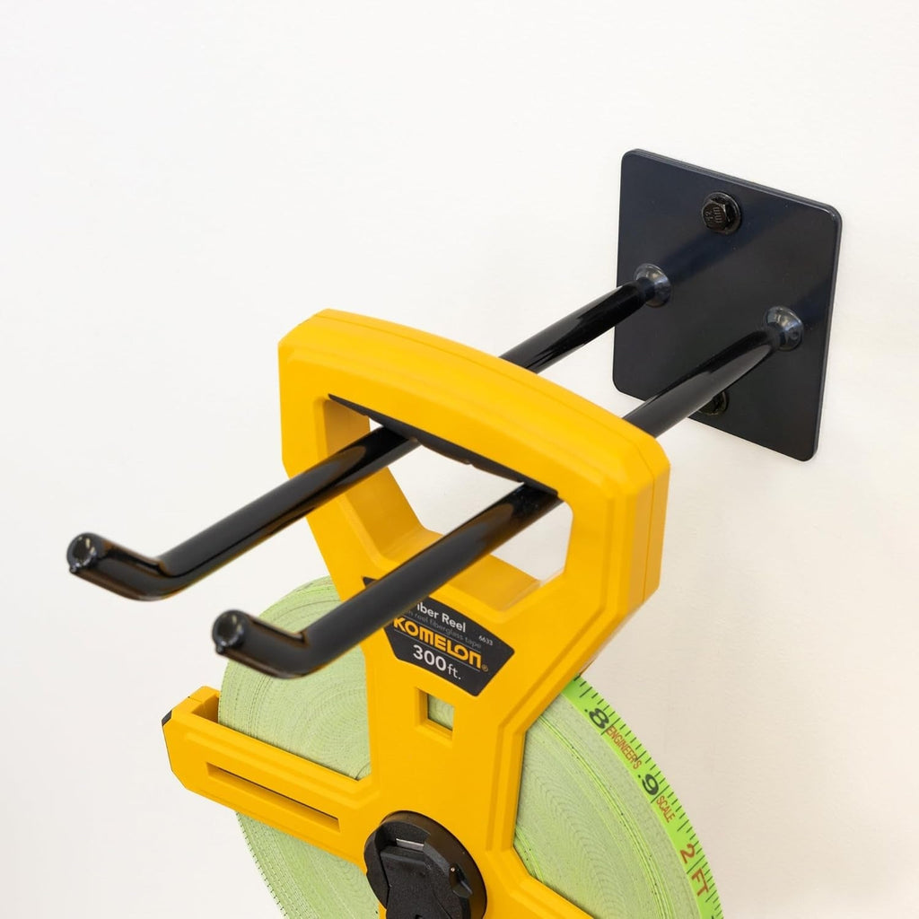 A yellow measuring tape reel hangs from the Bedrock Garage Wall Hook | Heavy-Duty | Deep Water, which features a double design and is securely mounted with a square base plate on a white wall.