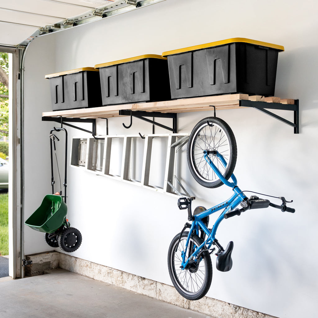 A garage wall displays 2' x 8' DIY Wall Shelf Brackets | Heavy Duty Garage Wall Shelf with four black bins with yellow lids, a metal ladder below, plus a blue bicycle and green spreader hanging on hooks.