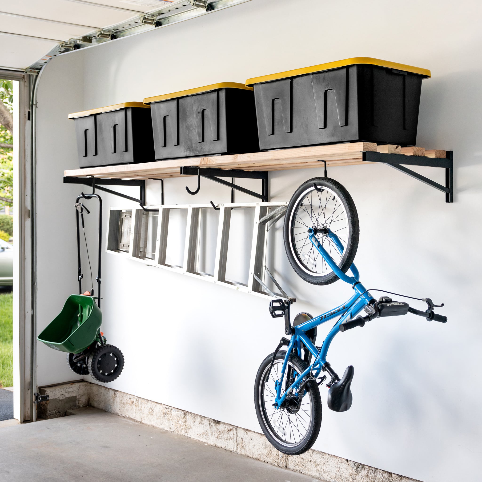 A garage wall displays 2' x 8' DIY Wall Shelf Brackets | Heavy Duty Garage Wall Shelf with four black bins with yellow lids, a metal ladder below, plus a blue bicycle and green spreader hanging on hooks.