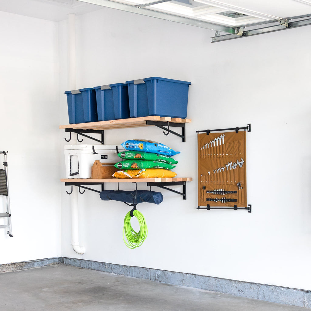Two wooden shelves on a white garage wall hold blue bins, gardening supplies, a lantern, and a toolbox. Below hangs a coiled green hose, and nearby is the Pivot Panel Tool Organizer in Harvest Tan, neatly storing hand tools.