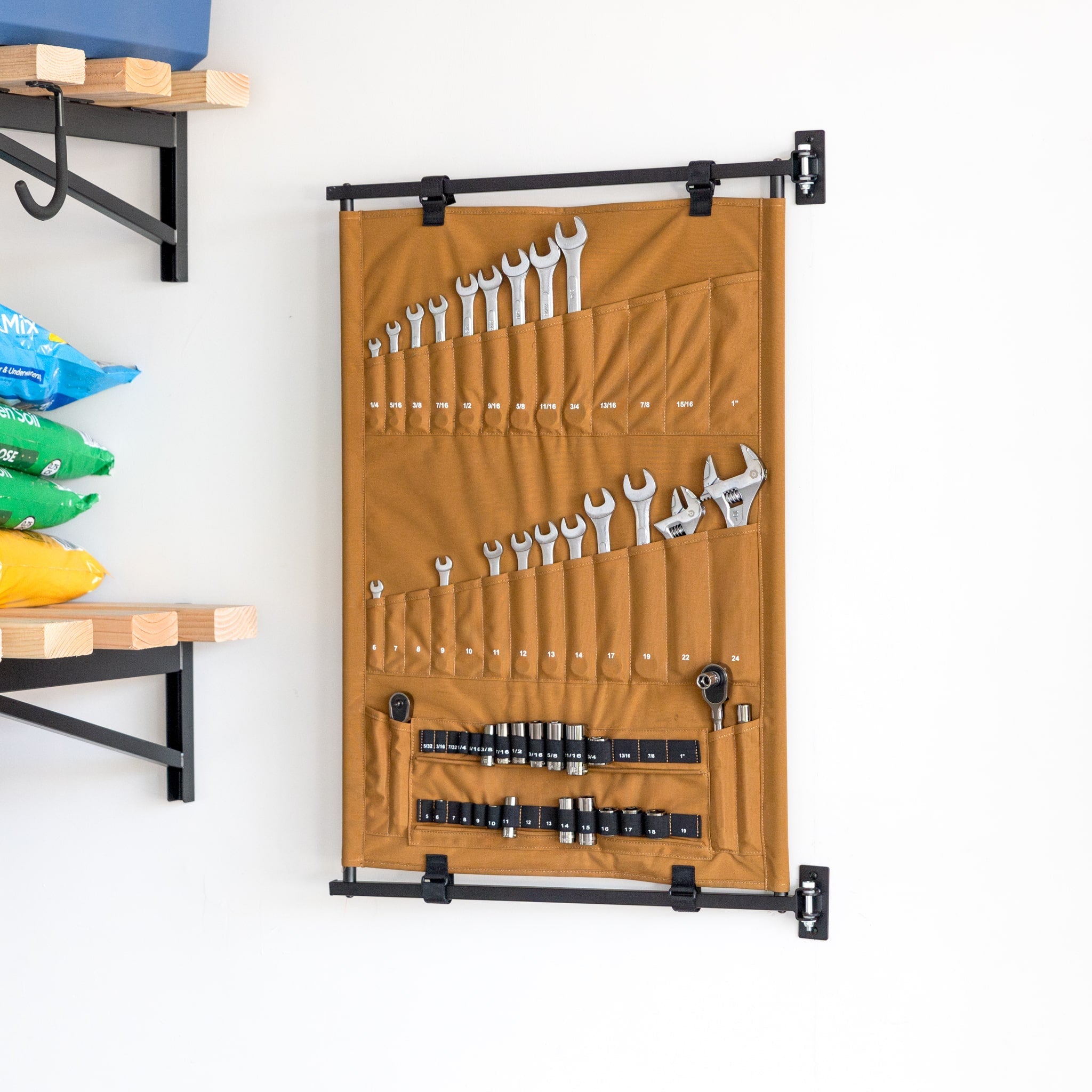 The Pivot Panel Tool Organizer in Harvest Tan hangs on a wall, holding wrenches and adjustable spanners in pockets, with shelves of colorful bags visible on the left.