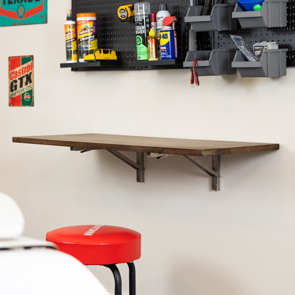 The Wakefield Foldable Workbench, featuring a dark wood tabletop and stainless hardware, is mounted beneath a pegboard with tools and bottles. A red stool sits below, with automotive and cleaning supplies organized nearby in the workspace.