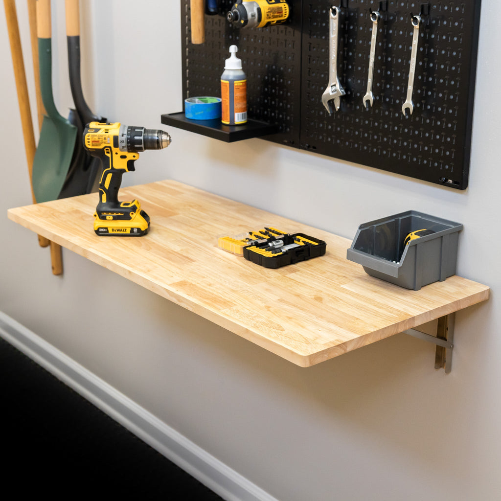 The Wakefield Foldable Workbench holds a cordless drill, tool organizer, and plastic bin. Above it, a black pegboard displays tools, while garden tools and a folding wall table are neatly arranged on the left.