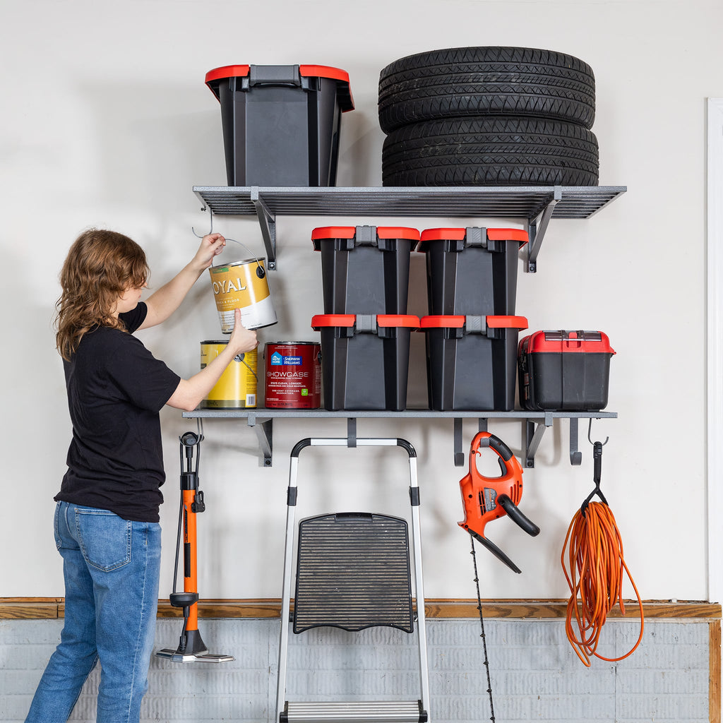 A person organizes paint cans on 2' x 4' Bristol Garage Wall Mount Shelves (2 Pack, Hammered Gray), which also hold black bins, tires, and tools. Hanging below are a leaf blower and garden tool. A step stool stands nearby.