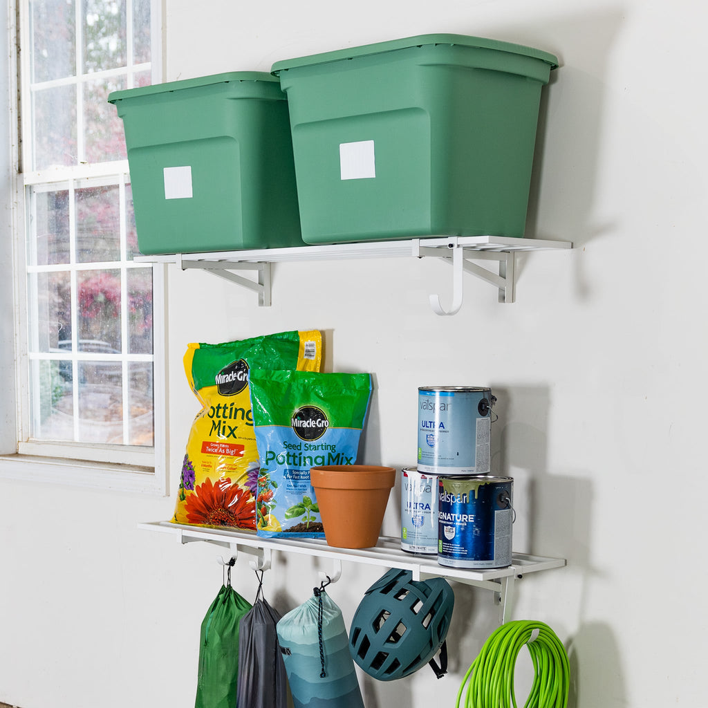 Two green bins sit on a white 1' x 4' Bristol Garage Wall Mount Shelf above another shelf with potting mix, paint cans, a flowerpot, and spray bottle. Hooks below hold bags, a bike helmet, and a green hose. A window is to the left.