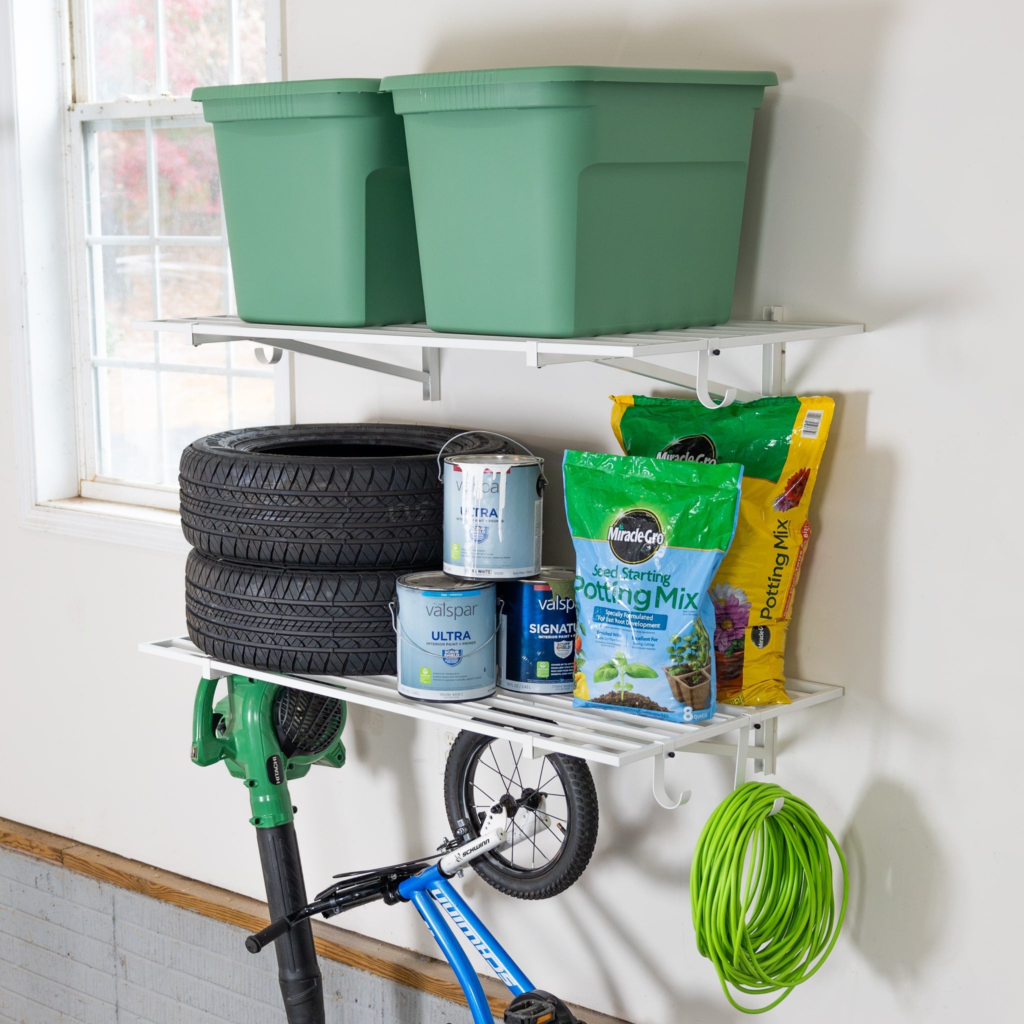 Two 2' x 4' Bristol Garage Wall Mount Shelves (2 Pack, White) hold green bins, tires, paint cans, potting mix bags, a green hose and trimmer. A blue bike rests below, while storage hooks provide extra organization in the bright garage.