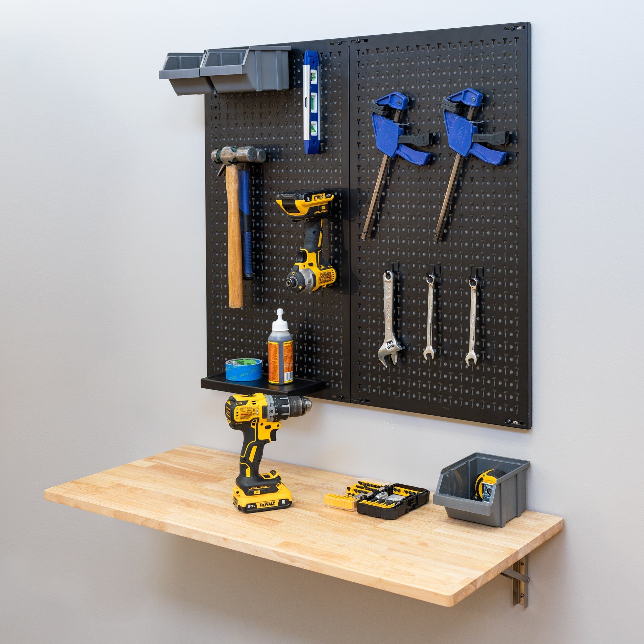 The Folding Workbench and Pegboard Workshop Set features a wall-mounted black pegboard for organizing tools like hammers, clamps, wrenches, and a level. The wooden workbench below holds a cordless drill, drill bits, and a bin with extra tools.
